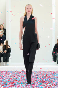 Model walks a runway wearing a black halter dress with side panels and pockets as pink-blue confetti falls around her on a white backdrop.