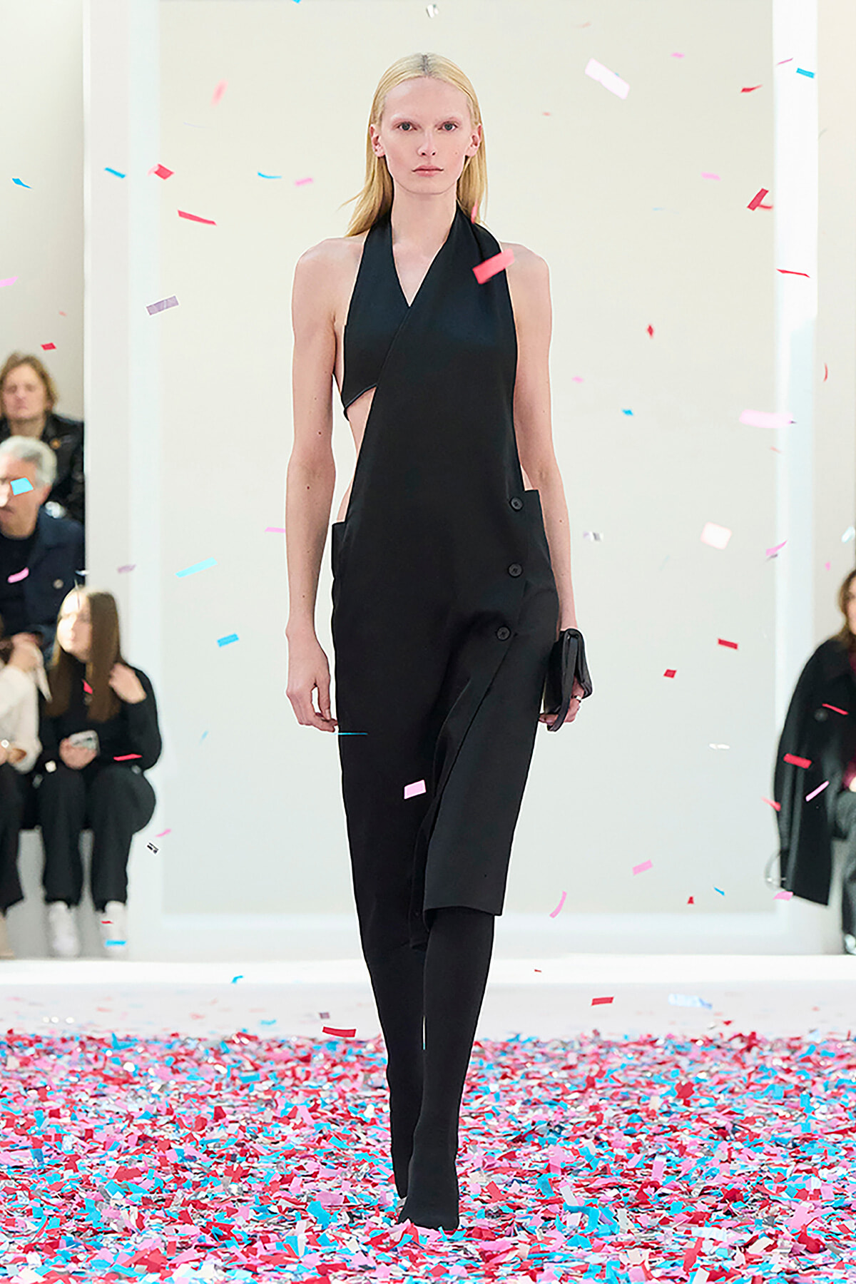 Model walks a runway wearing a black halter dress with side panels and pockets as pink-blue confetti falls around her on a white backdrop.