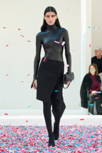 Model walking a fashion runway in a black shiny turtleneck and wrap skirt amid red and blue confetti.