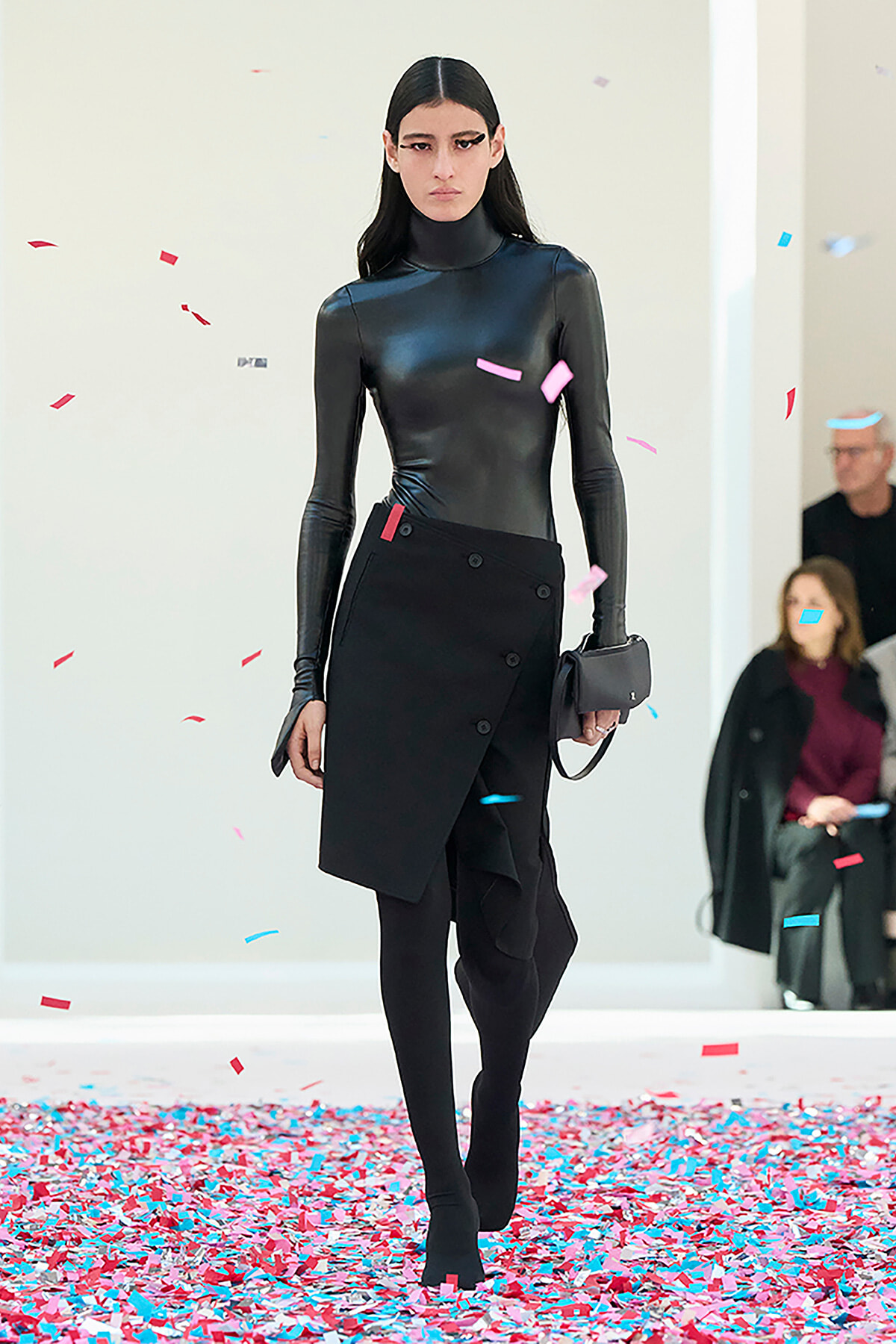 Model walking a fashion runway in a black shiny turtleneck and wrap skirt amid red and blue confetti.