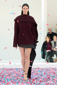 Runway model wearing a burgundy oversized top with a black mini skirt, with colorful confetti falling around her.