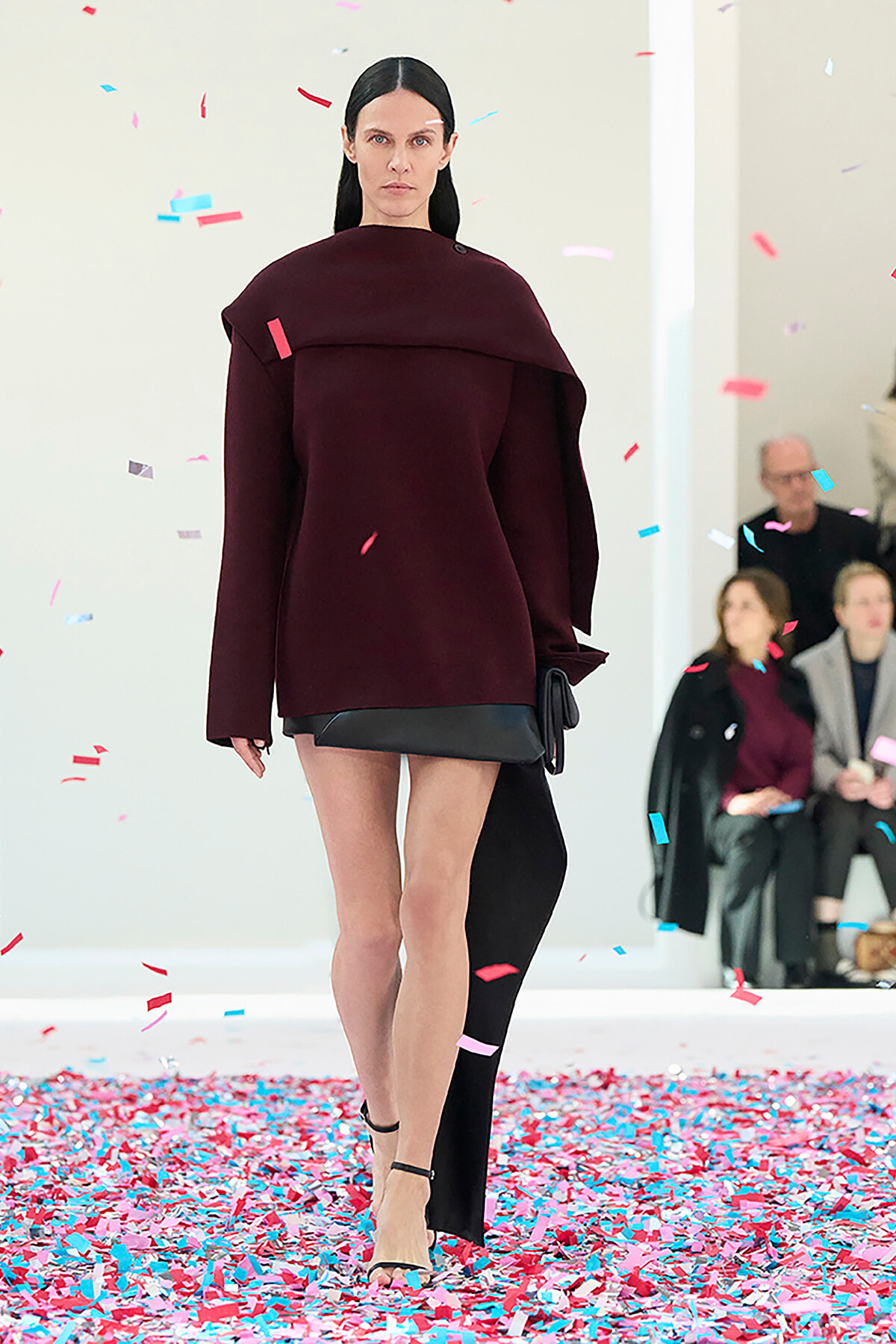 Runway model wearing a burgundy oversized top with a black mini skirt, with colorful confetti falling around her.
