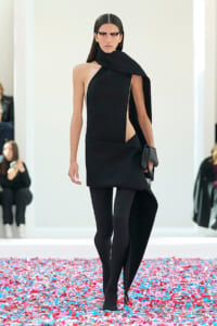 Model in a black asymmetric dress with a draped scarf, black tights, and a clutch walking a confetti-covered runway.