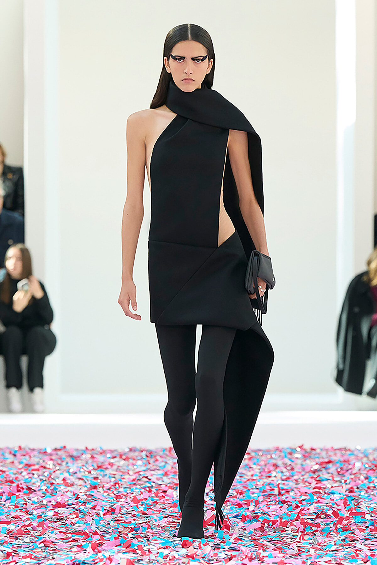 Model in a black asymmetric dress with a draped scarf, black tights, and a clutch walking a confetti-covered runway.