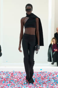 Model in a black asymmetrical crop top, high-waisted pants, and a draped cape walking the runway with futuristic sunglasses and a small clutch. Confetti covers the floor in pinks and blues.