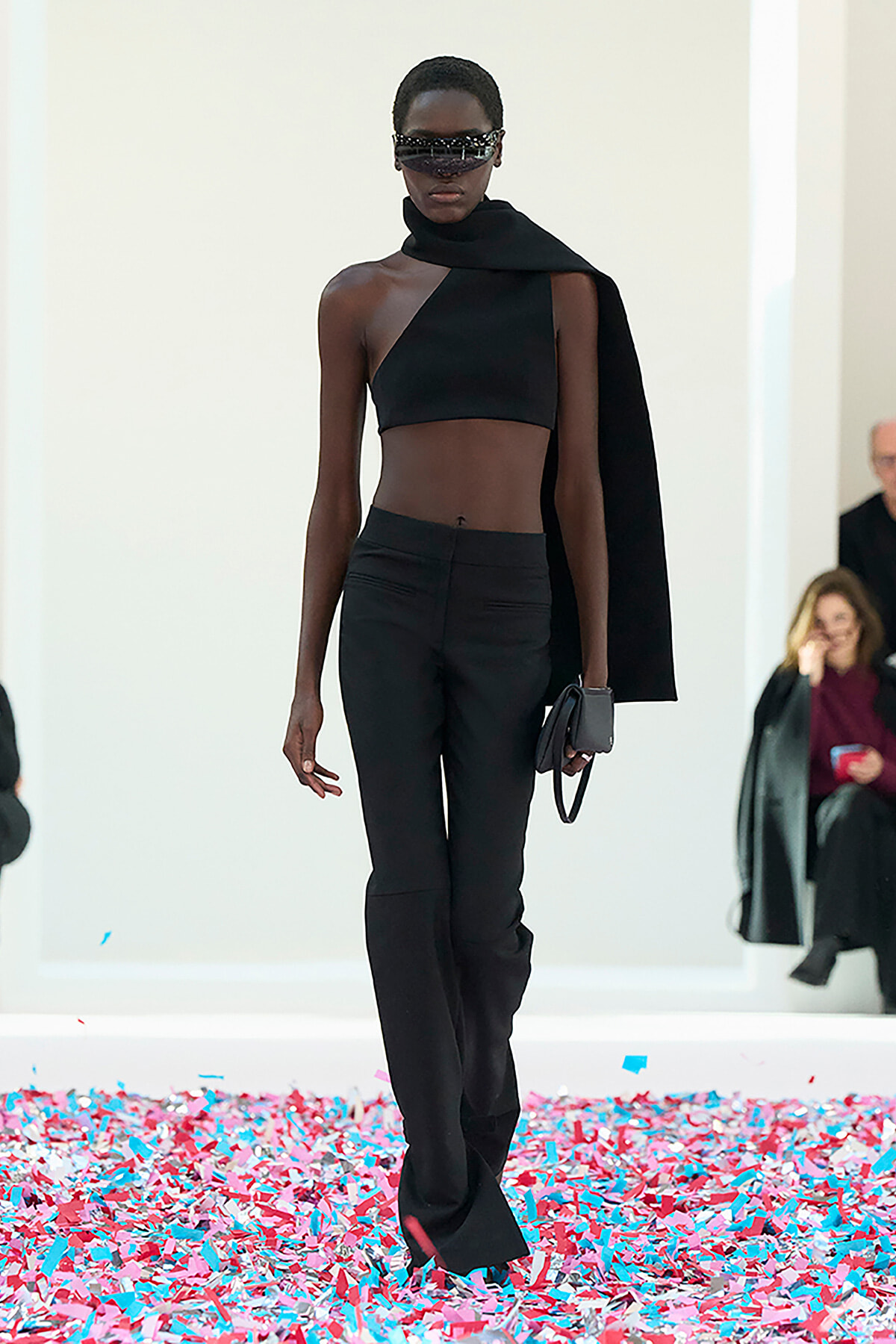 Model in a black asymmetrical crop top, high-waisted pants, and a draped cape walking the runway with futuristic sunglasses and a small clutch. Confetti covers the floor in pinks and blues.