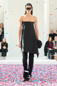 Model on a runway wearing a black strapless dress with an asymmetric hem and matching black pants, surrounded by falling confetti.