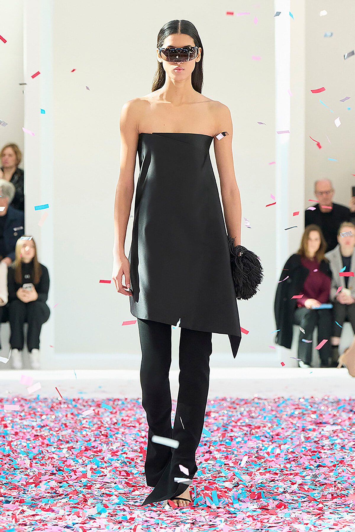 Model on a runway wearing a black strapless dress with an asymmetric hem and matching black pants, surrounded by falling confetti.