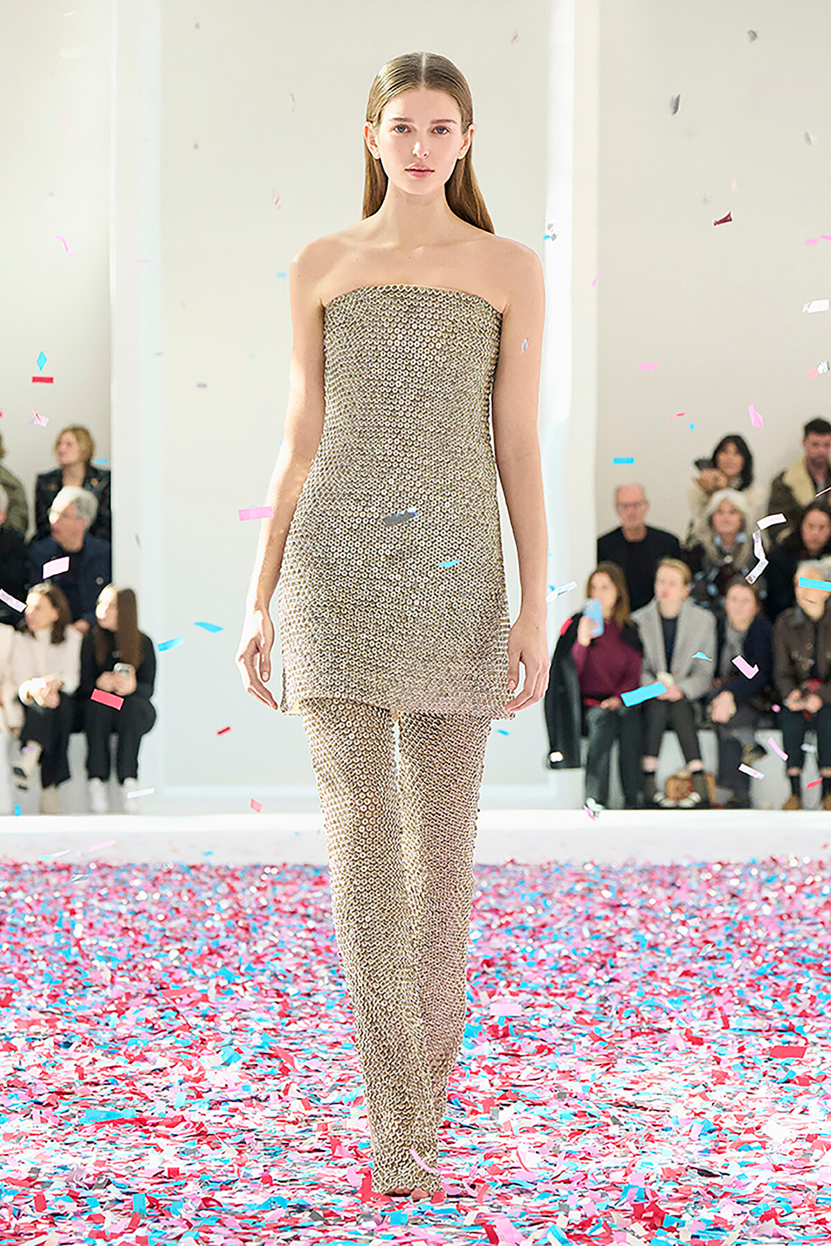 Runway model in a gold beaded strapless jumpsuit walking as colorful confetti falls around her.
