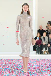 Model walks a fashion show wearing a beige geometric-print turtleneck midi dress with a pale pink clutch, confetti on the floor and audience in the background.