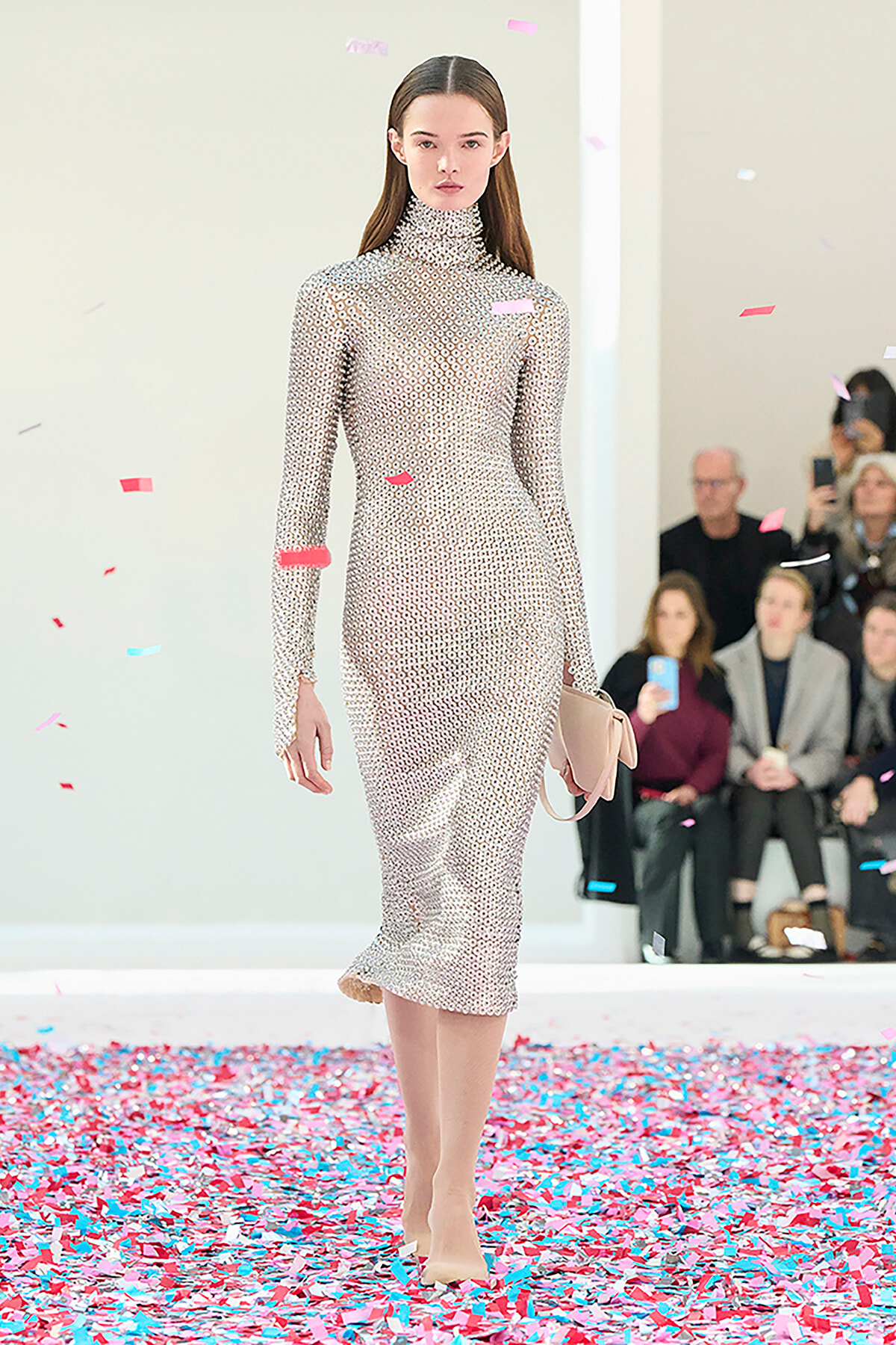 Model walks a fashion show wearing a beige geometric-print turtleneck midi dress with a pale pink clutch, confetti on the floor and audience in the background.