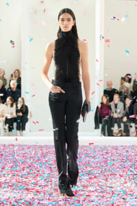 Model walking the runway in a black fringed top and black pants, holding a clutch, as confetti falls around her.