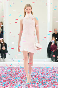 Model walks a runway in a white feathered dress with a matching clutch as pink and blue confetti falls around her.
