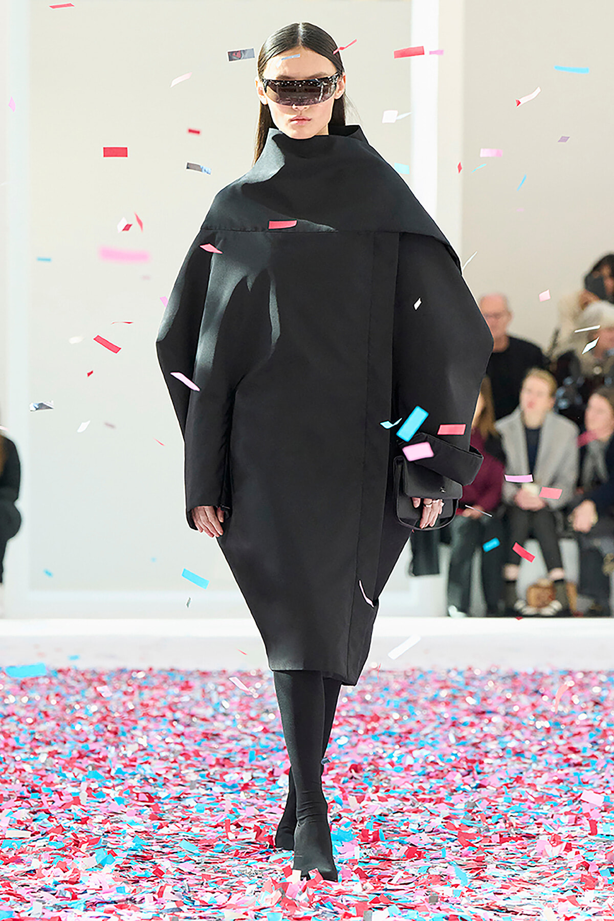 Model walks the runway in a black oversized coat and futuristic visor sunglasses as colorful confetti falls around her.