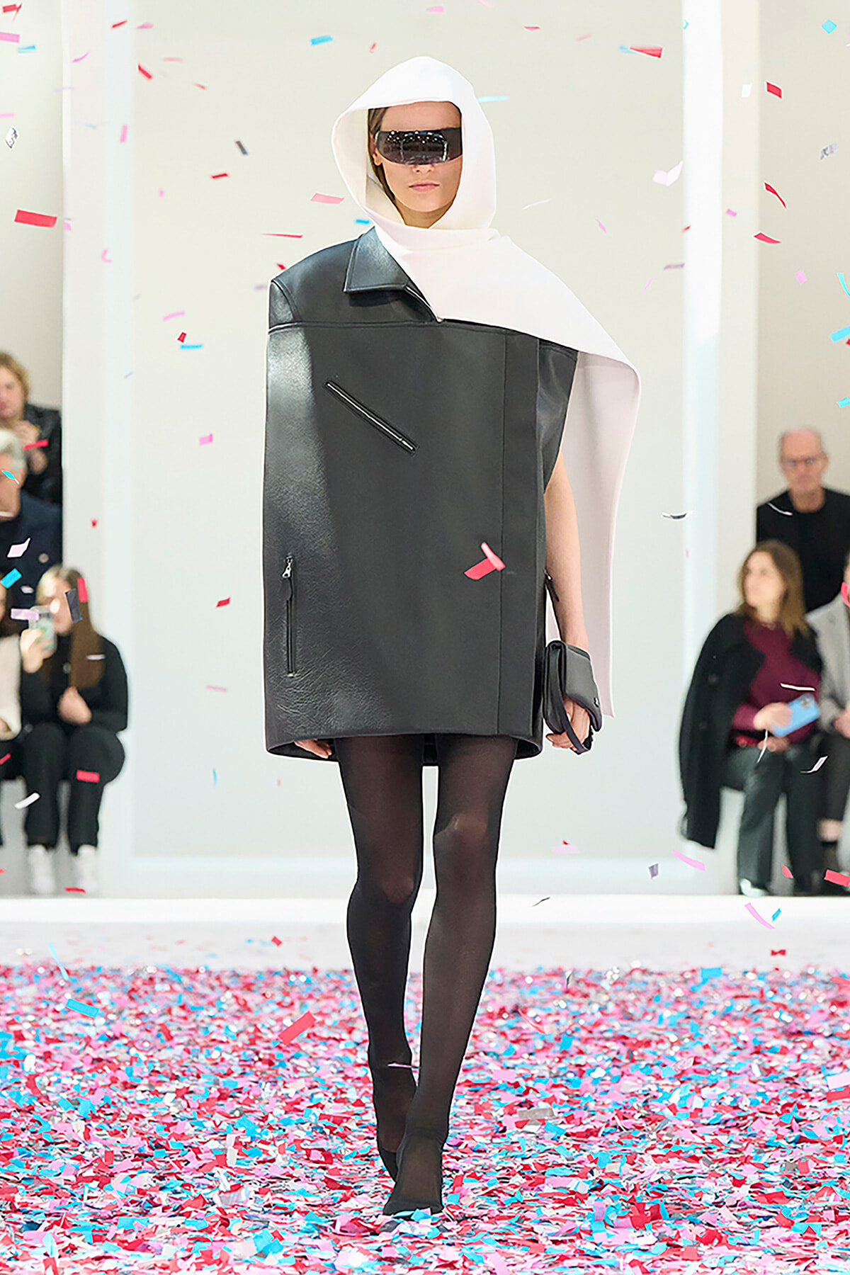 Model wearing futuristic black leather oversized vest with a white hooded scarf and dark sunglasses on a confetti-strewn runway.
