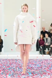 Model walks the runway in a minimalist white coat-dress as colorful confetti falls around her.