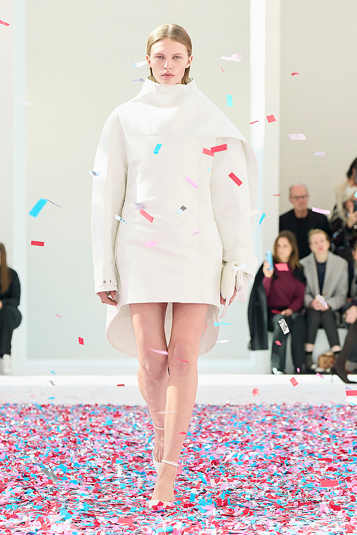 Model walks the runway in a minimalist white coat-dress as colorful confetti falls around her.