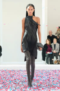 Model walking a fashion show in a black one-shoulder dress and thigh-high boots, carrying a black clutch amid colorful confetti on the runway