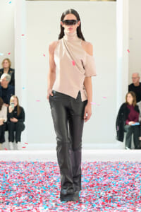 Model on a fashion runway wearing a beige asymmetrical top and black leather pants, with futuristic sunglasses; colorful confetti on the floor.