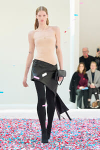 Model walks a runway in a strapless beige bodysuit and an asymmetric black skirt, black tights, and a small clutch, with colorful confetti on the floor and audience in the background.