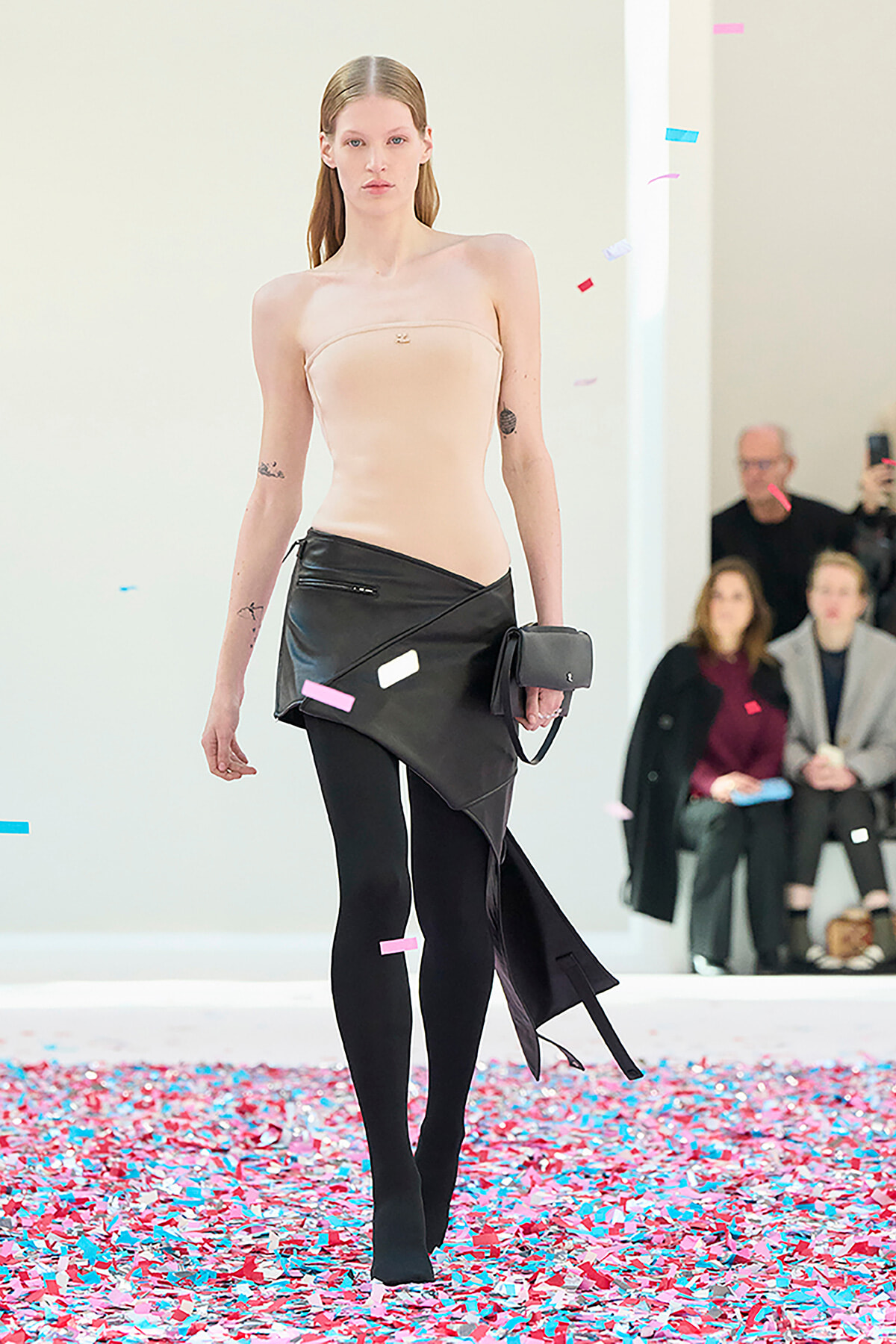Model walks a runway in a strapless beige bodysuit and an asymmetric black skirt, black tights, and a small clutch, with colorful confetti on the floor and audience in the background.