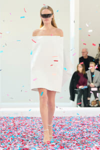 Model walks a runway in an off-the-shoulder white dress and futuristic black visor, with colorful confetti falling around.