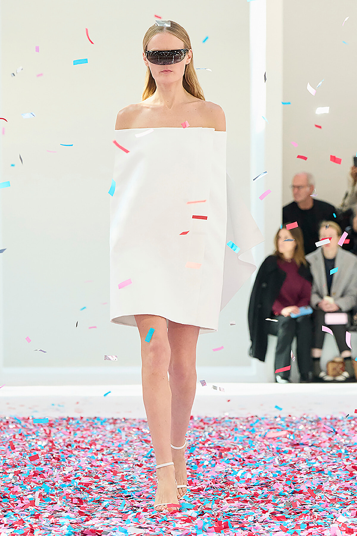 Model walks a runway in an off-the-shoulder white dress and futuristic black visor, with colorful confetti falling around.