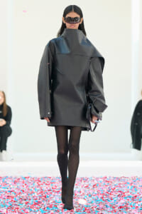 Model walks a runway in an oversized black coat, black tights, and bold sunglasses; confetti on the floor around her.
