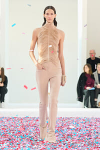 Model walks the runway in a sleeveless beige feathered top and matching pale pants, surrounded by falling confetti.