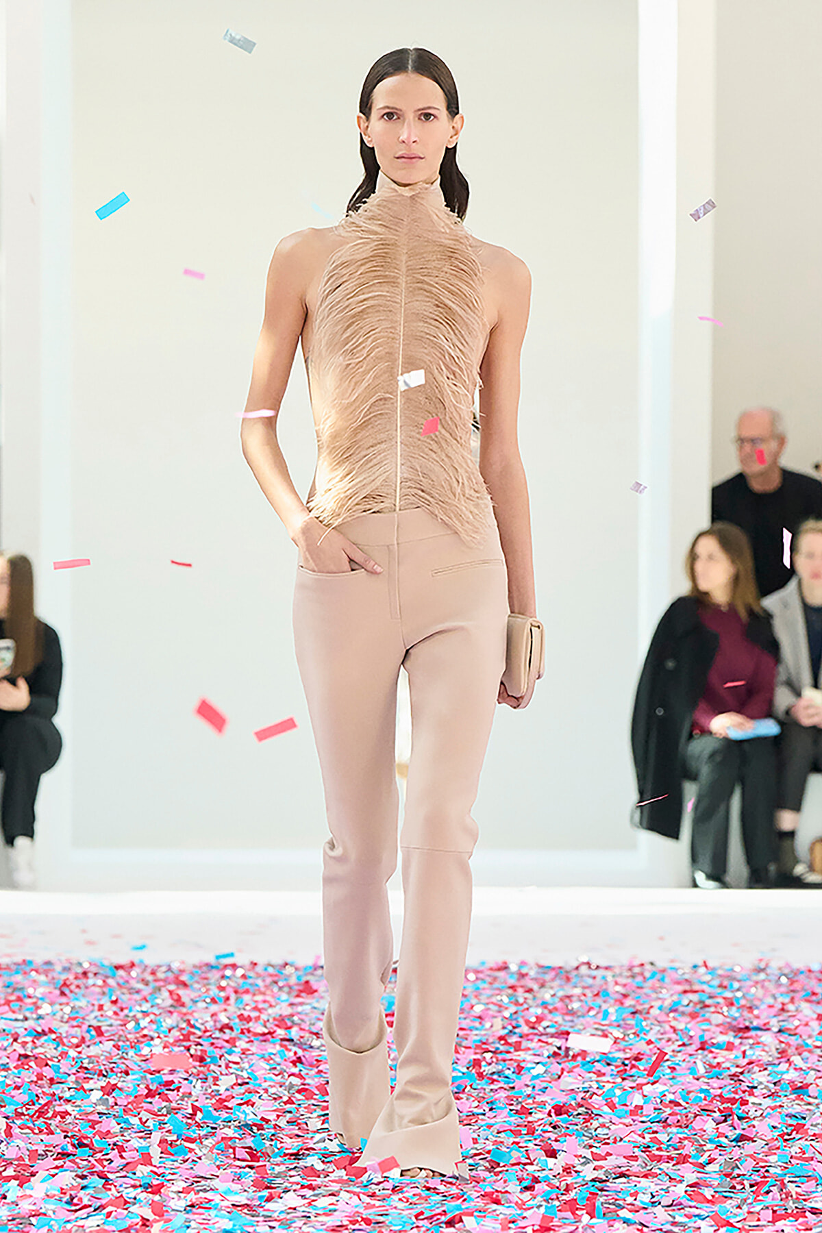 Model walks the runway in a sleeveless beige feathered top and matching pale pants, surrounded by falling confetti.