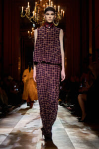 Model walks a fashion show in a burgundy geometric-print sleeveless top and matching wide-leg pants, under chandeliers in a grand venue.