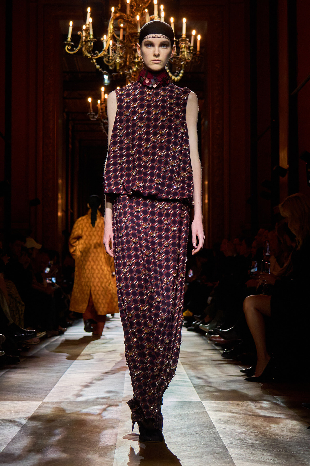Model walks a fashion show in a burgundy geometric-print sleeveless top and matching wide-leg pants, under chandeliers in a grand venue.