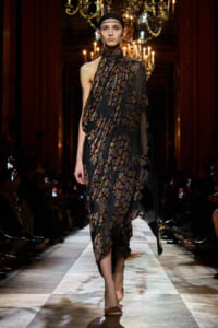 Model on a runway wearing a sheer, dark floral-printed dress with asymmetrical draping and a headband, chandeliers visible in the background