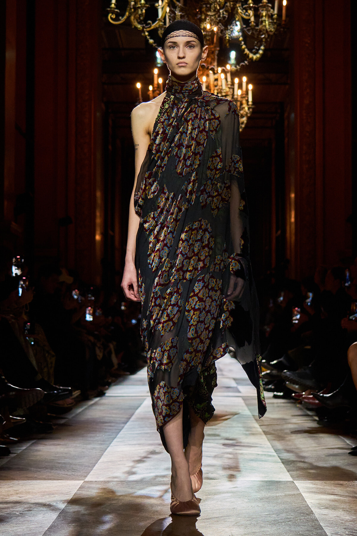 Model on a runway wearing a sheer, dark floral-printed dress with asymmetrical draping and a headband, chandeliers visible in the background