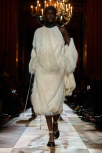 Model on a runway wearing a large, fluffy white shaggy dress with a black headwrap, chandeliers in the background.