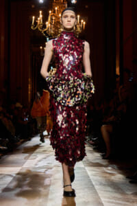 Model walks the runway in a burgundy, scale-like sequin dress with a high neckline and headband, illuminated by chandeliers in a luxurious venue.