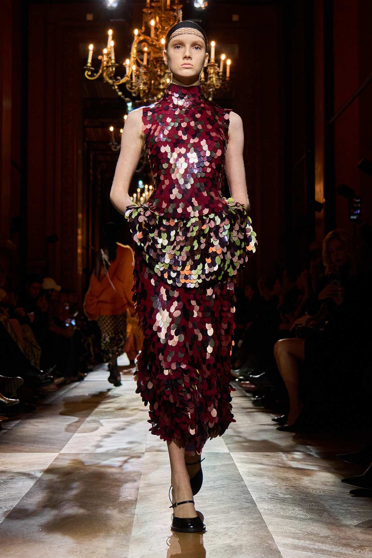 Model walks the runway in a burgundy, scale-like sequin dress with a high neckline and headband, illuminated by chandeliers in a luxurious venue.