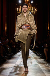 Runway model in a chunky beige patterned knit cape with a wide belt and dark patterned tights on a lit catwalk.