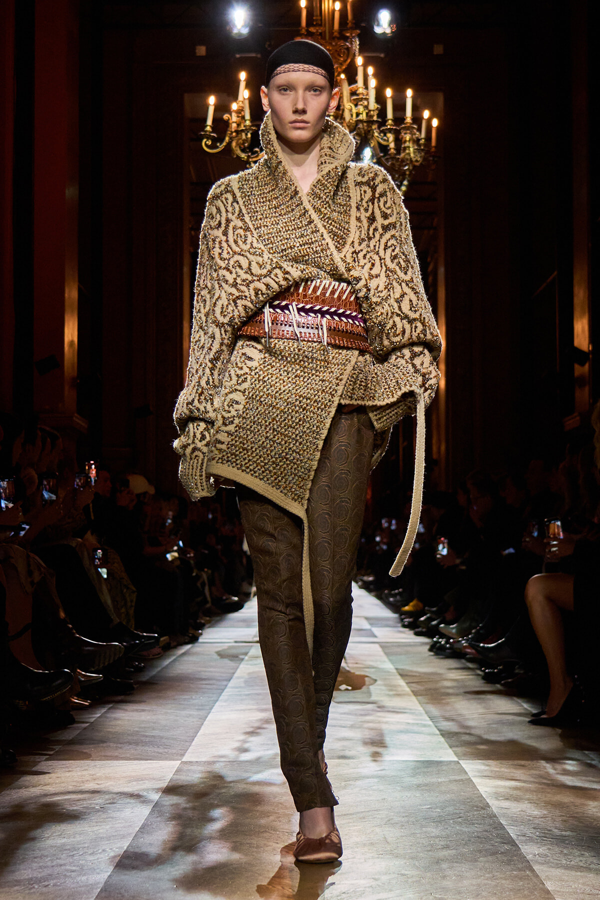 Runway model in a chunky beige patterned knit cape with a wide belt and dark patterned tights on a lit catwalk.