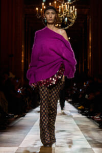 Model walking a runway in a bright magenta oversized knit sweater draped over one shoulder with patterned wide-leg pants and a dark headband, chandeliers in the background.