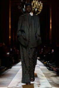 Runway model in a dark dotted oversized suit with embellished pants, hands in pockets, chandeliers in the background.