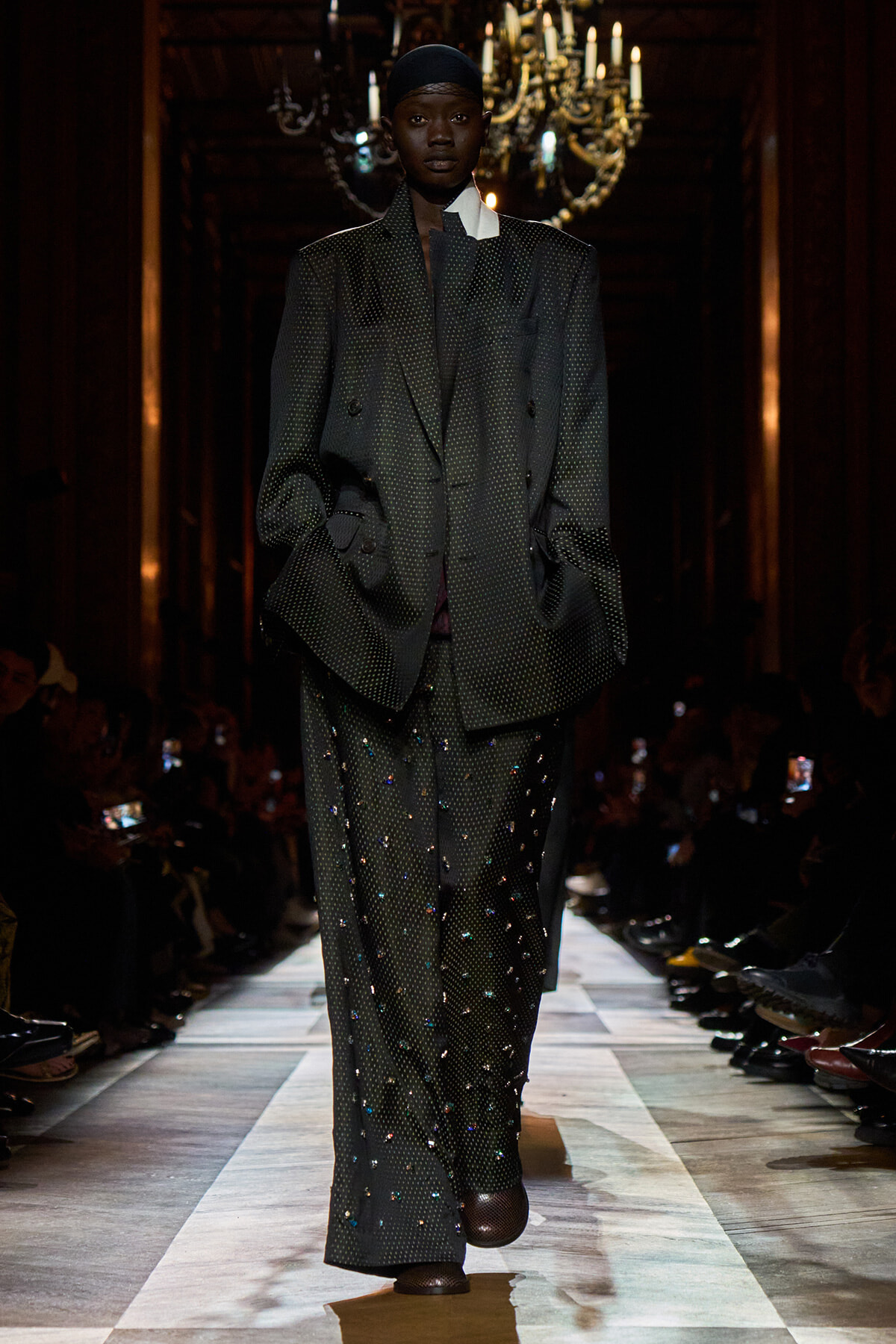 Runway model in a dark dotted oversized suit with embellished pants, hands in pockets, chandeliers in the background.