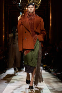Model walks the runway in a rust oversized coat over a chunky knit turtleneck and layered patterned skirts.