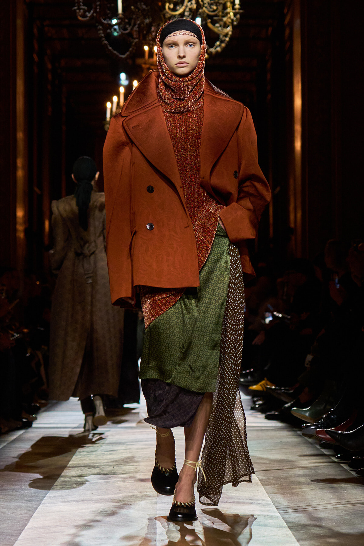 Model walks the runway in a rust oversized coat over a chunky knit turtleneck and layered patterned skirts.
