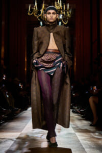 Model on a runway wears a long brown brocade coat opened at the chest, layered with patterned scarf and multicolor pants, head covered with a black cap.