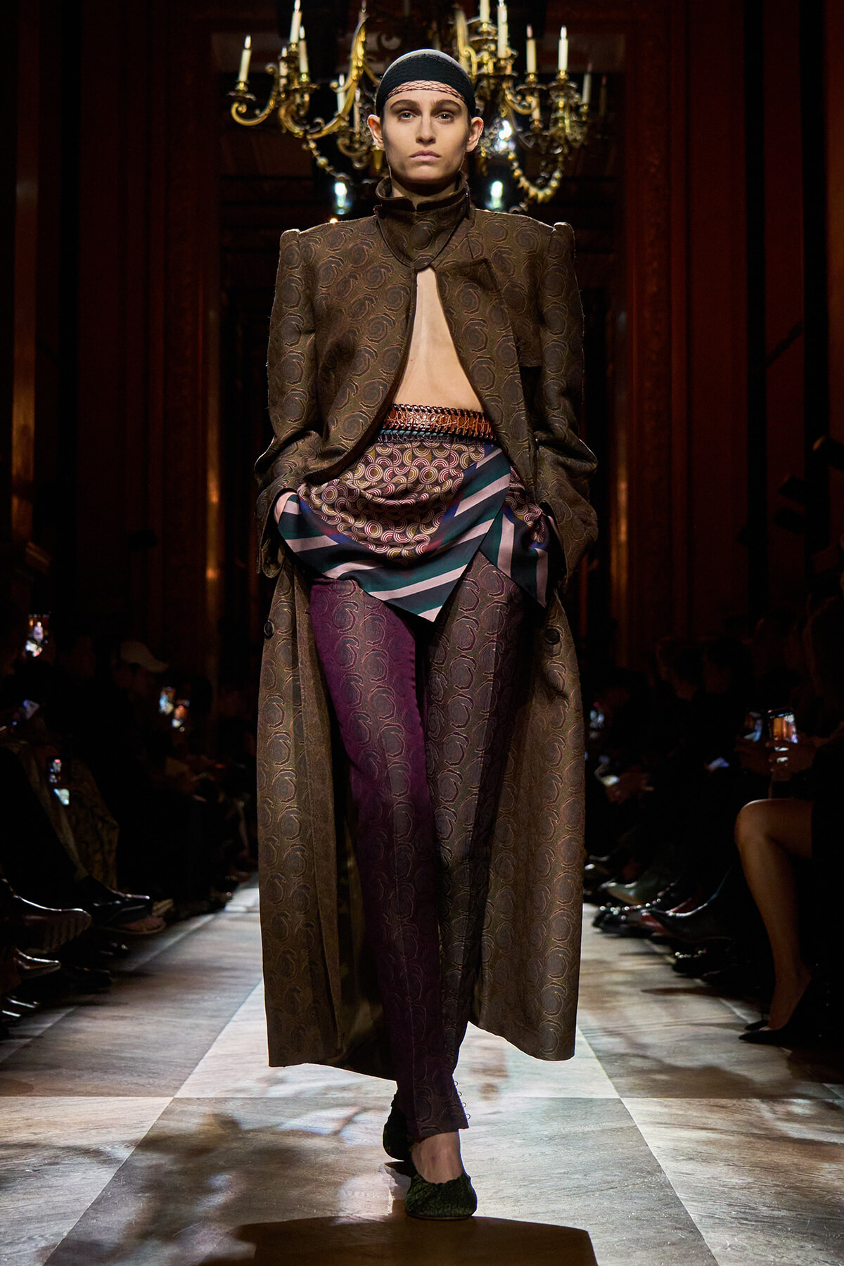 Model on a runway wears a long brown brocade coat opened at the chest, layered with patterned scarf and multicolor pants, head covered with a black cap.