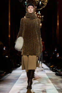 Model walking runway in a sparkling bronze knit poncho with a high cowl hood, beige skirt, tall brown boots, and a white fluffy handbag.