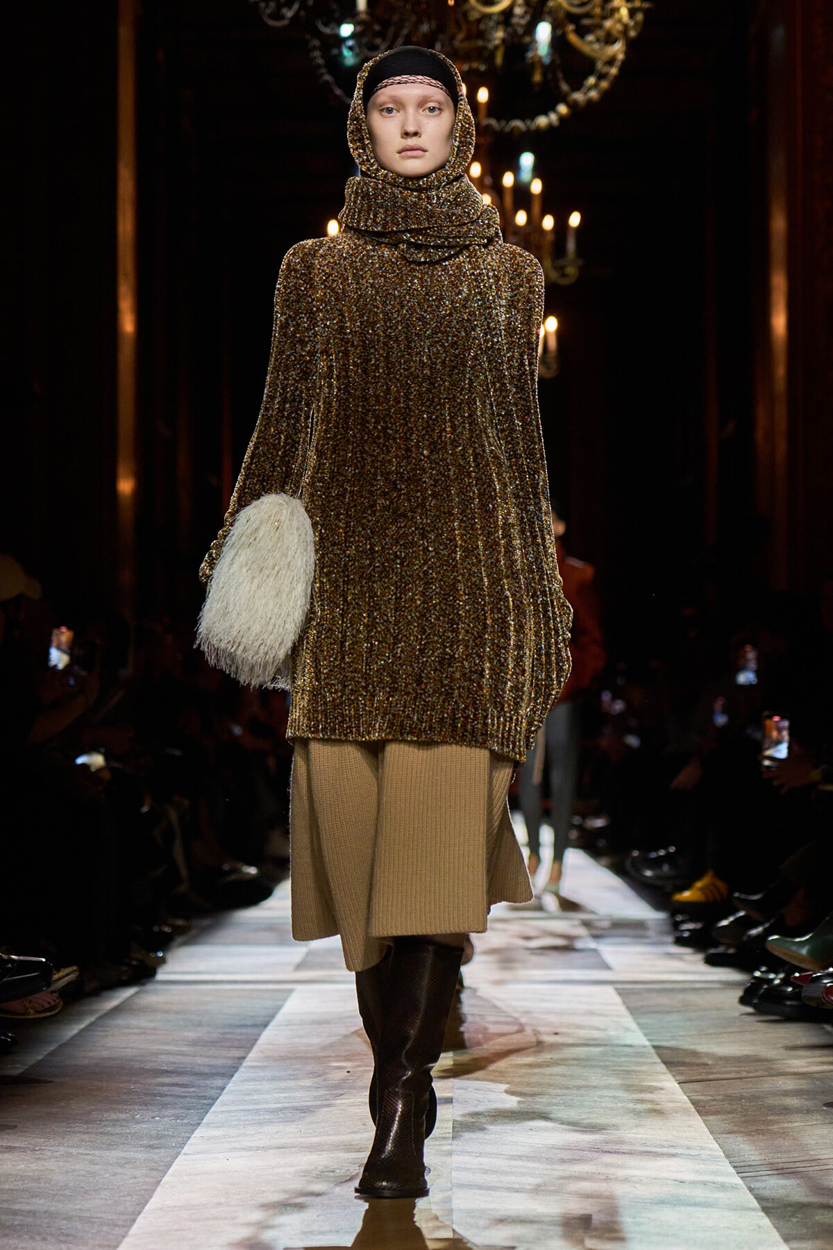 Model walking runway in a sparkling bronze knit poncho with a high cowl hood, beige skirt, tall brown boots, and a white fluffy handbag.