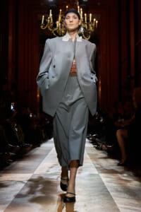Model walking the runway in an oversized gray suit with a tailored blazer and wide-legged pants, paired with a cropped top and black headband, under chandeliers on the catwalk.