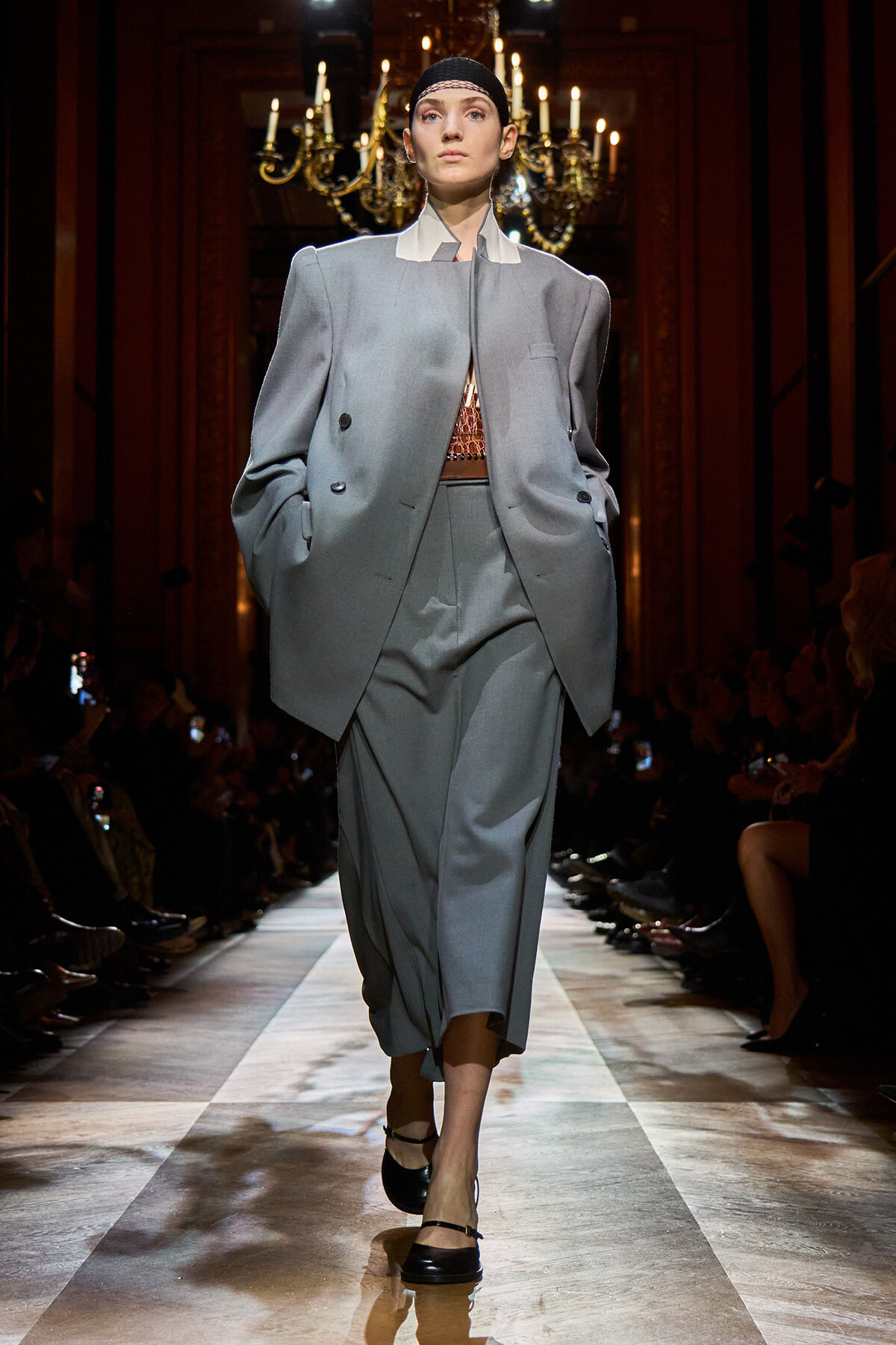Model walking the runway in an oversized gray suit with a tailored blazer and wide-legged pants, paired with a cropped top and black headband, under chandeliers on the catwalk.